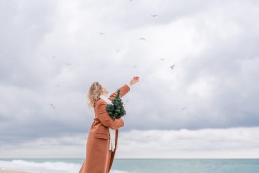 Blond woman Christmas sea. Christmas portrait of a happy woman walking along the beach and holding a Christmas tree on her shoulder. She is wearing a brown coat and a white suit