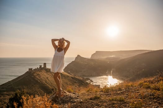 A blonde woman stands on a hill overlooking the ocean. She is wearing a white dress and she is enjoying the view