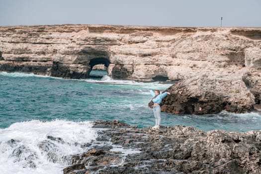 A woman in a blue jacket stands on a rock above a cliff above the sea and looks at the raging ocean. Girl traveler rests, thinks, dreams, enjoys nature. Peace and calm landscape, windy weather