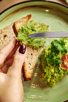 Elegant female hands spreading homemade guacamole on slice of toasted artisanal bread on background of ceramic plate. Light Mexican style snack