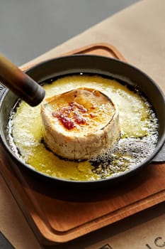 Culinary scene capturing preparation of wheel of Camembert cheese, lightly burned with cooking torch for golden melted surface in cast iron skillet placed on wooden board