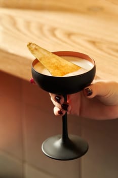 Female hand elegantly holding black stemmed glass with sweetish light alcoholic cocktail garnished with spicy caramelized pear slice against blurred background of wooden bar counter