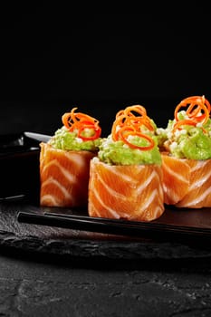 Close-up of salmon wrapped sushi rolls topped with crushed avocado and wasabi garnished with airy rice balls and vegetable shavings served on slate black board with chopsticks