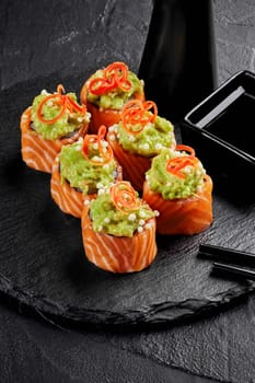 Salmon sushi rolls adorned with zesty avocado mix, bell pepper shavings and crispy rice balls, arranged on slate board with soy sauce and black chopsticks