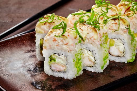 Close-up view of delicious uramaki sushi rolls topped with green tobiko, crab and creamy sauce garnished with scallion shavings on rustic ceramic plate