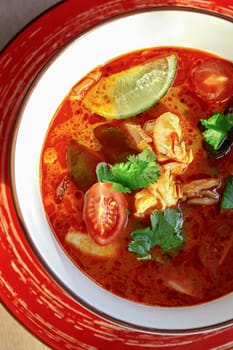 Close-up view of rich piquant seafood Tom Yum soup with shrimp, squid, shiitake mushrooms, tomatoes, and lime, garnished with fresh coriander on red ceramic plate background. Thai food concept