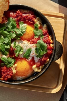 Delicious spicy shakshuka of two eggs in cast iron pan with rich tomato sauce and hummus, served with Greek yogurt and white bread toast garnished with fresh greens