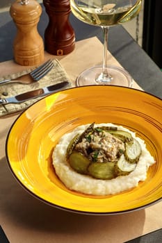 Elegant dinner setup featuring creamy beef stroganoff on mashed potatoes with sliced pickles served on yellow plate, accompanied by glass of white wine