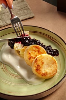 Female hands slicing browned fluffy syrniki served on ceramic plate with berry topping and delicate yoghurt cream. Traditional sweet dish of Ukrainian cuisine