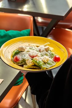 Customer holding Caesar salad with baked chicken fillet, fresh greens, crispy croutons, and cherry tomatoes sprinkled with grated parmesan, in vibrant yellow plate at cafe