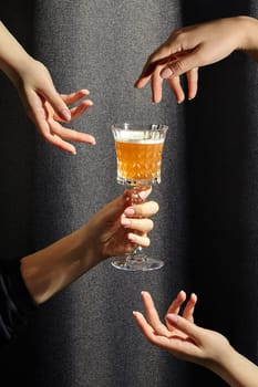 Female hands gracefully hovering around crystal glass of whiskey sour against dark textured fabric background with shadow. Concept of convivial spirit of sharing beloved cocktail