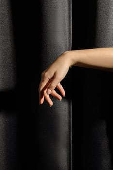 Graceful female hand in soft natural pose contrasting with texture of black curtain in dramatic lighting. Minimalist scene evoking themes of elegance, solitude, subtle interplay of light and darkness