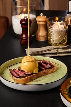 Perfectly cooked beef steak slices on toasted bread with poached egg topped with velvety hollandaise sauce, presented on ceramic plate in intimate dining setting
