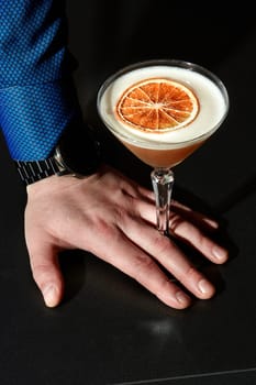 Elegant gentlemans hand leaning against black surface, holding glass of almond sour cocktail with frothy top adorned with dried grapefruit slice