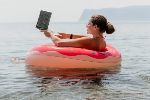 Woman freelancer works on laptop swimming in sea on pink inflatable ring. Pretty lady typing on computer while floating in the sea on inflatable donut at sunset. Freelance, remote work on vacation