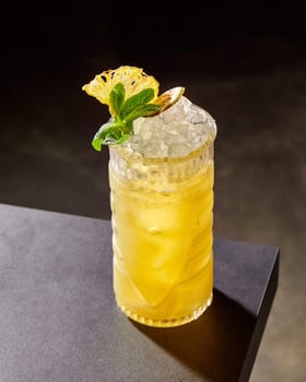 Sweetish tiki cocktail, tropical rum drink with pineapple juice and crushed ice garnished with fresh mint leaves and dried fruit slices, served in tall glass on dark background