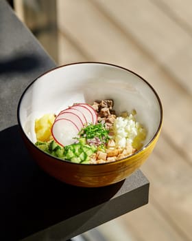 Bowl basking in sunlight showcasing vibrant finely diced ingredients for okroshka, refreshing Russian summer soup, with crisp radishes, cucumber, potatoes, eggs, meat and fresh herbs