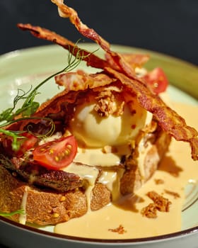 Delicious bruschetta with slices of juicy veal steak and poached egg dressed with creamy hollandaise sauce, topped with crispy onions and fried bacon garnished with greens and cherry tomatoes