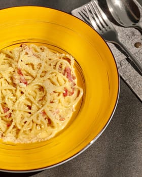 Classic Italian rich creamy spaghetti carbonara with bits of pancetta, garnished with grated parmesan, presented in striking yellow bowl on table in sunlight. Authentic cuisine concept