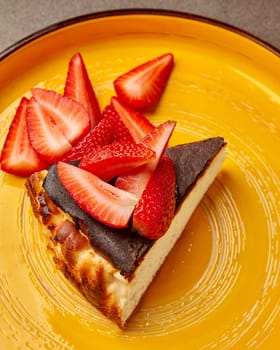 Slice of tempting soft cheesecake with browned crust served on vibrant yellow ceramic plate with succulent fresh ripe strawberries. Sweet indulgence concept