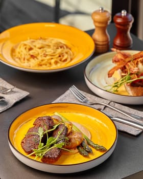 Warm inviting meal presented on vibrant yellow plates including pork steak with baked potatoes and asparagus, creamy pasta and bacon-topped bruschetta in background