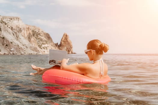 Woman freelancer works on laptop swimming in sea on pink inflatable ring. Pretty lady typing on computer while floating in the sea on inflatable donut at sunset. Freelance, remote work on vacation