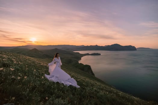 A woman in a white dress is standing on a hill overlooking a body of water. The sky is a mix of orange and purple hues, creating a serene and romantic atmosphere. The woman is enjoying the view