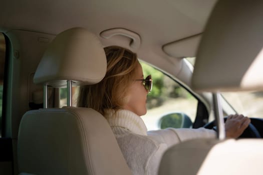 A blonde woman in a white sweater and jeans is driving. Happy woman sitting in a car with a white interior