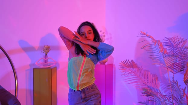 Attractive happy dancer looking at camera while smart woman moving at rhyme with neon light. Professional hispanic performer break dancing while wearing colorful cloth with diverse friend. Regalement.