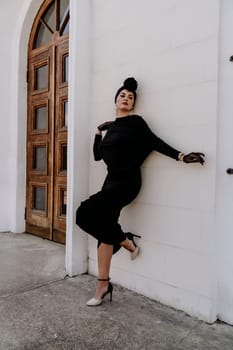 Stylish woman in the city. Fashion photo of a beautiful model in an elegant black dress posing against the backdrop of a building on a city street.