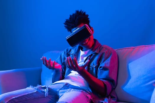 Young African American looking VR enjoy watching fantastic 3D pointing interesting object technology blue neon light virtual reality metaverse new world at living room sitting on sofa. Contrivance.