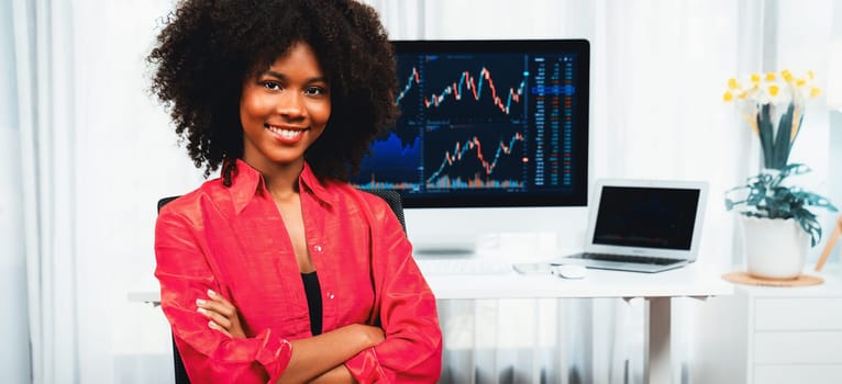 Analytical young African American businesswoman, a specialist in successful stock exchange trading, against dynamic data graph displaying marketing trend analysis on screen. Tastemaker.