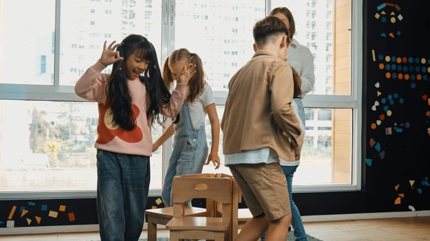 Diverse children playing musical chairs games while young smart beautiful teacher turn off music. Multicultural students running and sitting on chair in creative activity. Music lesson. Erudition.