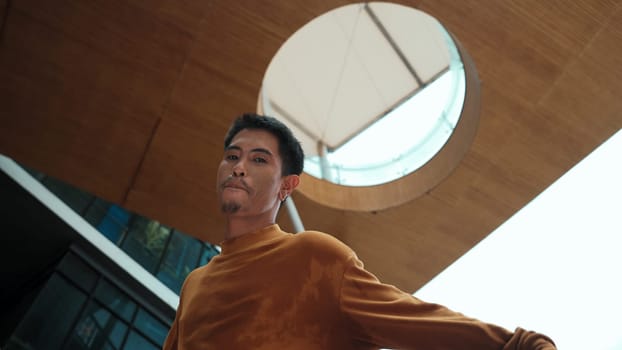 Hispanic dancer looking at camera while dancing at sky scrapper. Attractive handsome hipster moving his hands to hip hop music at modern mall with low angle camera. Outdoor sport 2024. Endeavor.