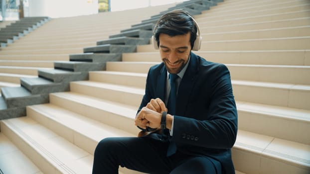 Smiling business man looking at smart watch while waiting for partner. Project manager listening music while sitting at stair surround by modern mall. Happy man wearing suit waiting at mall. Exultant.