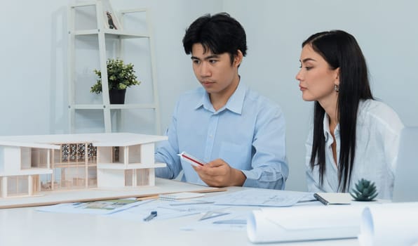 Professional male asian architect using ruler to measure house model length while young beautiful caucasian colleague using laptop to analyzed data on meeting table with house model. Immaculate.