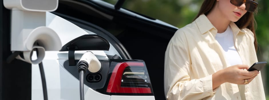 Young woman use smartphone to pay for electricity at public EV car charging station green city park. Modern environmental and sustainable urban lifestyle with EV vehicle. Panorama Expedient