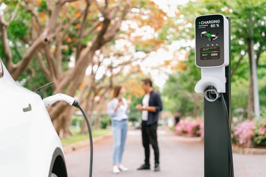 Focused EV car recharging battery on blurred background of lovey couple during autumnal road trip travel with electric vehicle recharging battery. Eco friendly travel on vacation during autumn. Exalt