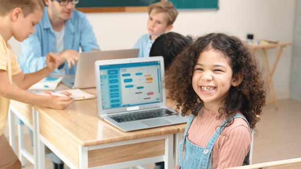 American student looking at camera and learning prompt in STEM technology class while group of diverse student and teacher doing mind mapping. Smart child learning coding software program. Pedagogy.