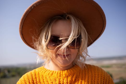 A woman wearing a brown hat and sunglasses is smiling. She is wearing a yellow sweater and has blonde hair