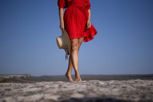 A woman in a red dress is standing on a rocky beach with a straw hat in her hand
