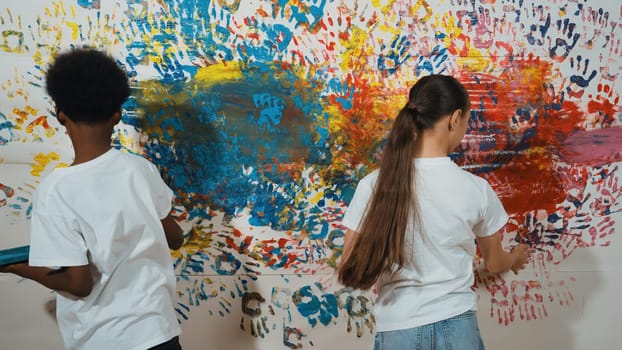 Back view of young diverse children paint colorful stained wall with hand. Attractive highschool student paint the wall with hand print while holding color palette. Creative activity. Edification.