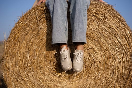 A person is sitting on a hay bale. The hay bale is round and has a lot of straw. The person is wearing white shoes