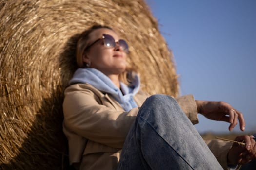 A woman is sitting in a hay bale, wearing sunglasses and a blue hoodie. She is enjoying the outdoors and taking a break from her day