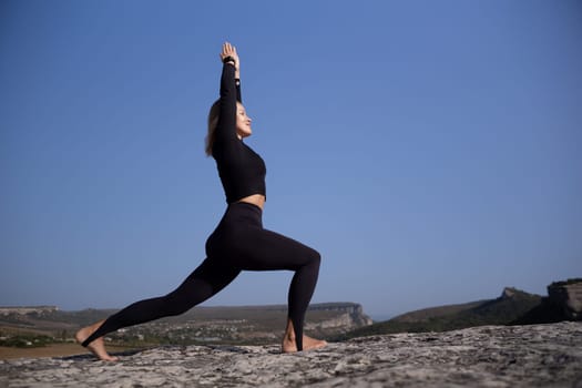 A woman is doing yoga on a rocky hillside. The sky is clear and blue, and the woman is wearing black clothing