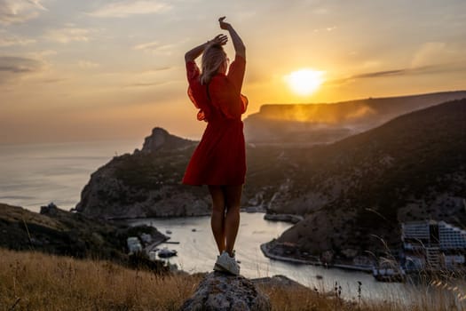 Happy woman standing with her back on the sunset in nature in summer with open hands posing with mountains on sunset, silhouette. Woman in the mountains red dress, eco friendly, summer landscape active rest.