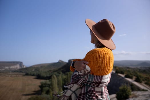 A woman wearing a brown hat and a yellow sweater is standing on a hillside, looking out at the landscape