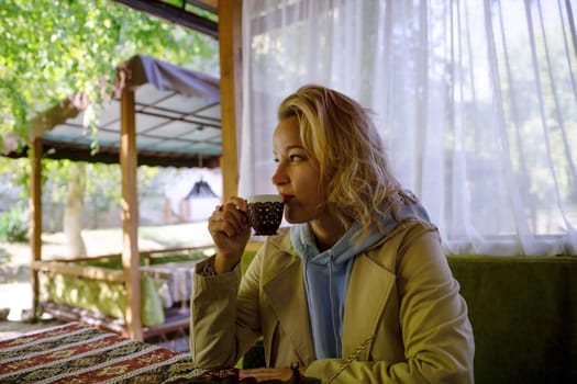 A woman is sitting at a table with a cup of coffee in front of her. She is wearing a tan jacket and a blue hoodie. The scene is set outdoors, with a bench and a canopy nearby