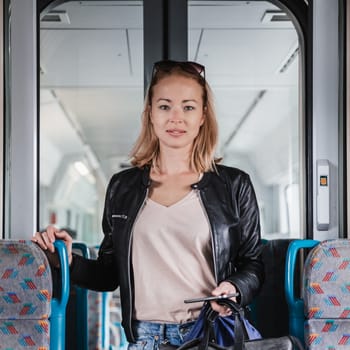 Young blond woman in jeans, shirt and leather jacket holding her smart phone and purse while riding modern speed train arriving to final train station stop. Travel and transportation