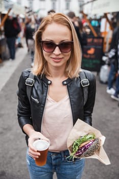 Beautiful young woman holding delicious organic salmon vegetarian burger and homebrewed IPA beer on open air beer an burger urban street food festival in Ljubljana, Slovenia